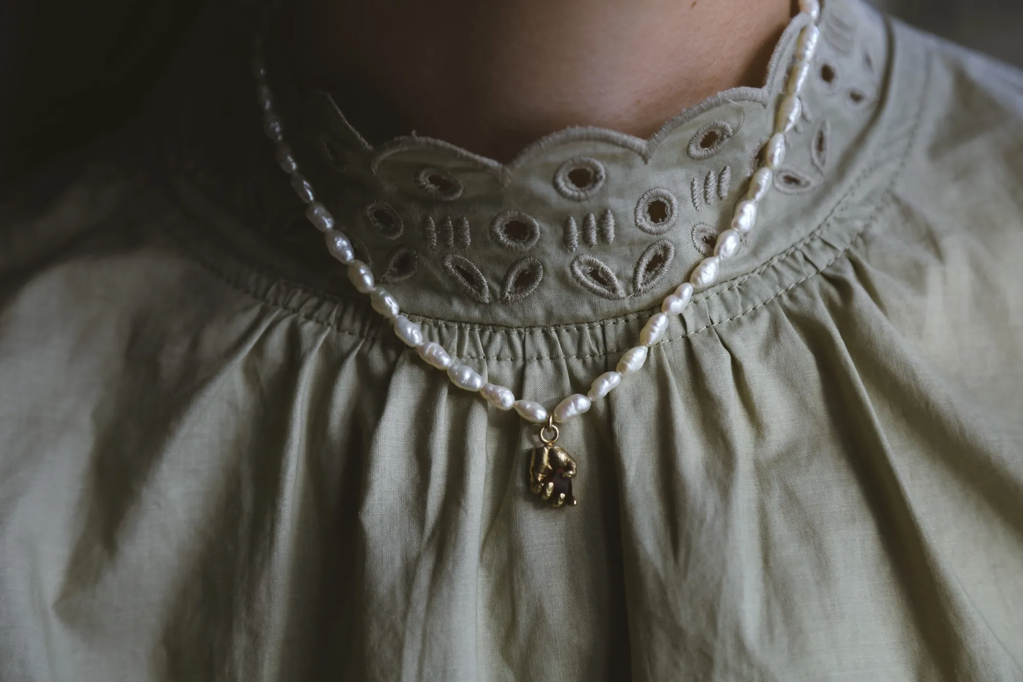 A freshwater pearl necklace with a small gold hand-and-garnet pendant, worn against a sage-green embroidered collar