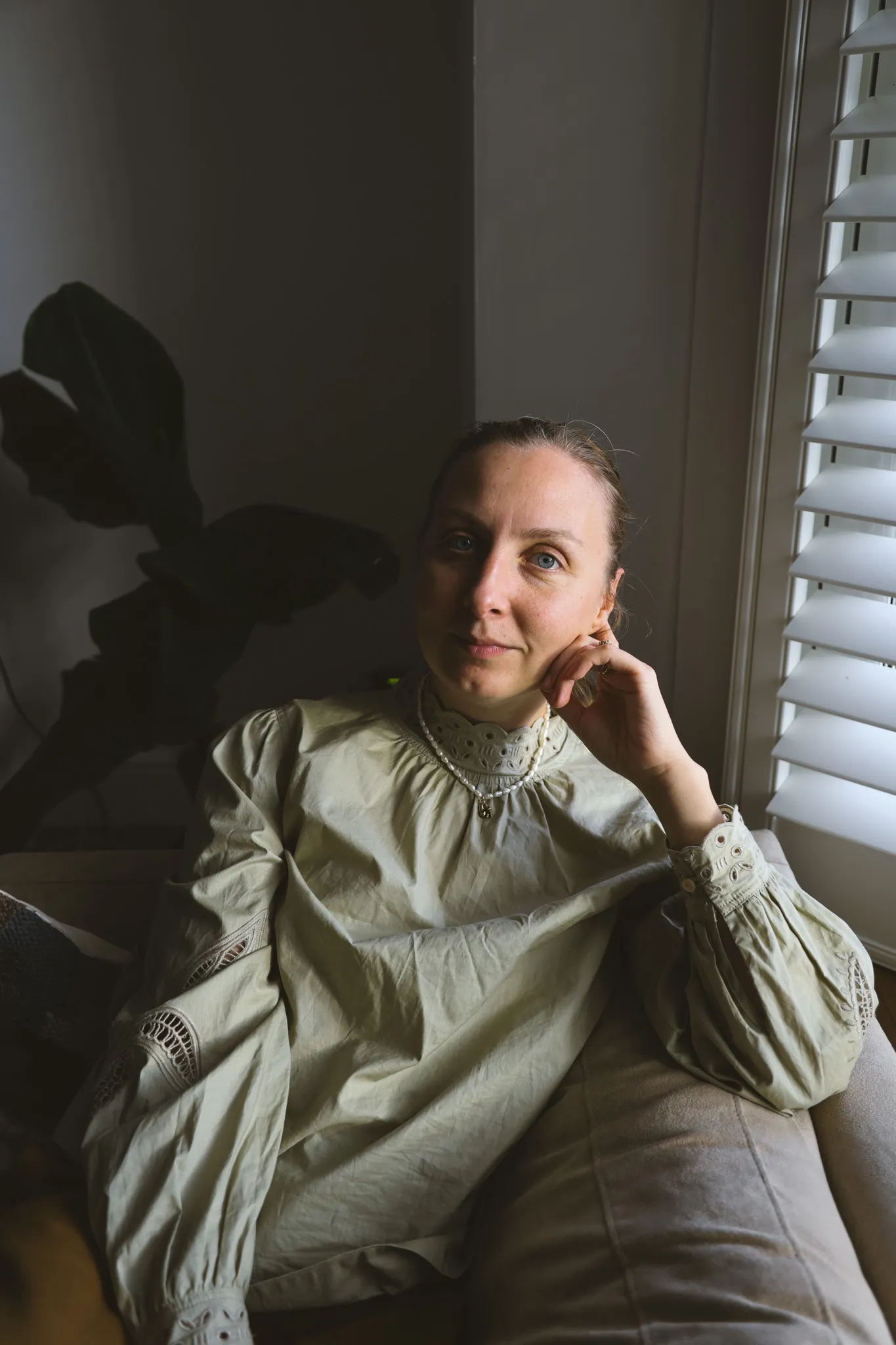 A woman in a sage-green embroidered blouse resting her cheek on her hand, wearing a freshwater pearl necklace with a small gold pendant, beside a window with white shutters