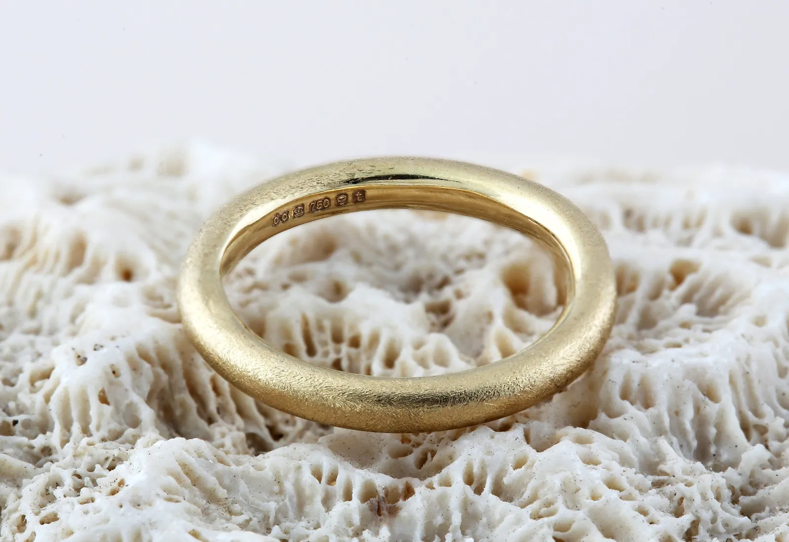 A brushed-texture gold wedding band resting on white coral, showing its hallmark inside the band